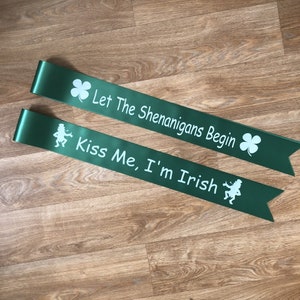 May include: Two green satin sashes with white text. One sash says "Let The Shenanigans Begin" with a shamrock. The other sash says "Kiss Me, I'm Irish" with a leprechaun.