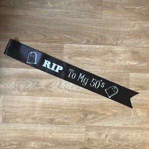 May include: Black satin sash with white text and tombstone graphics. The sash reads "RIP To My 50's". A fun accessory for a milestone birthday celebration.