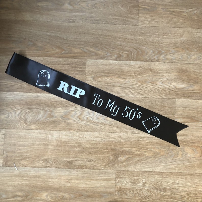May include: Black satin sash with white text and tombstone graphics. The sash reads "RIP To My 50's". A fun accessory for a milestone birthday celebration.