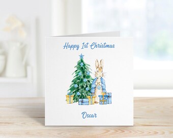 May include: A white card with a blue inscription that reads "Happy 1st Christmas". A watercolor illustration of a rabbit holding a blue ornament in front of a Christmas tree with blue ornaments and presents. The name "Oscar" is written in blue at the bottom of the card.