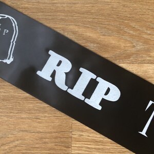 May include: Black satin sash with the text "RIP" in large, white, bold letters. A white tombstone graphic is visible on the left side. The sash is angled across a wooden surface.