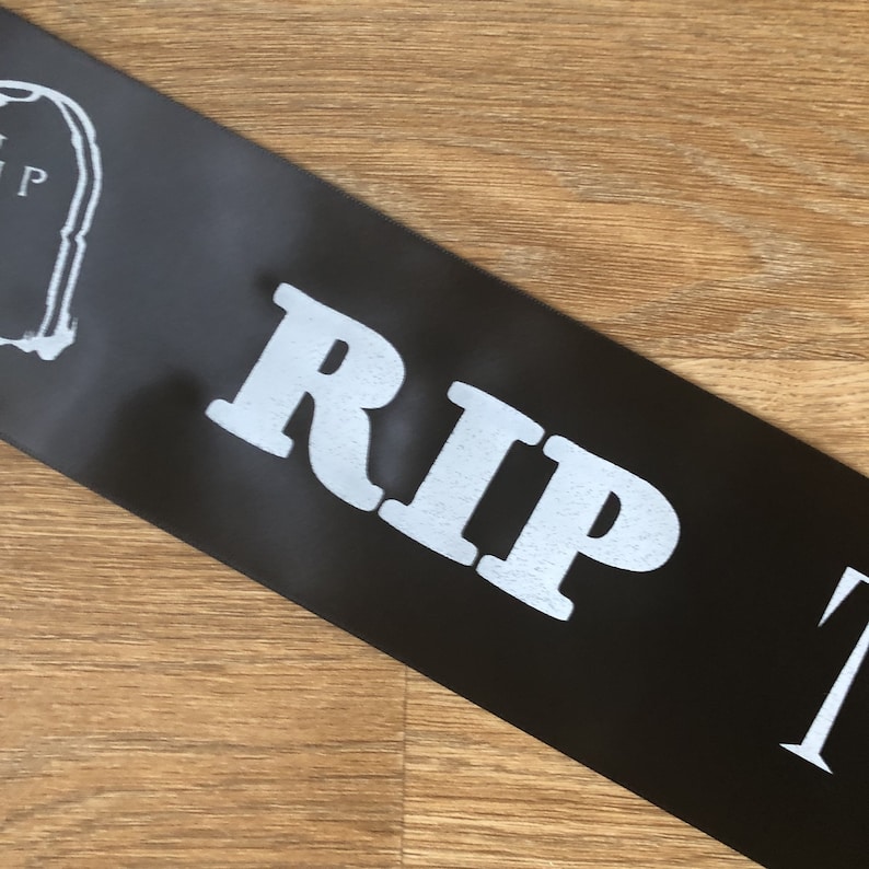 May include: Black satin sash with the text "RIP" in large, white, bold letters. A white tombstone graphic is visible on the left side. The sash is angled across a wooden surface.