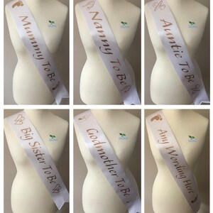 May include: Six white satin sashes with gold lettering. The sashes read "Mummy To Be", "Nanny To Be", "Auntie To Be", "Big Sister To Be", "Godmother To Be", and "Any Wording Here".
