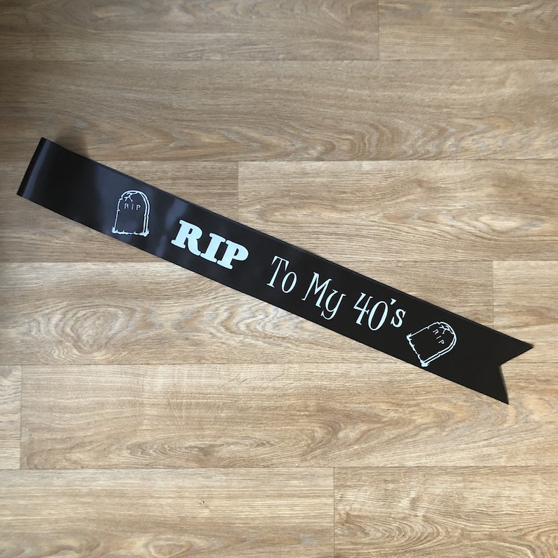 May include: Black satin sash with the text "RIP To My 40's" in white and light blue. The sash features a tombstone graphic at each end. A humorous accessory for a milestone birthday celebration.