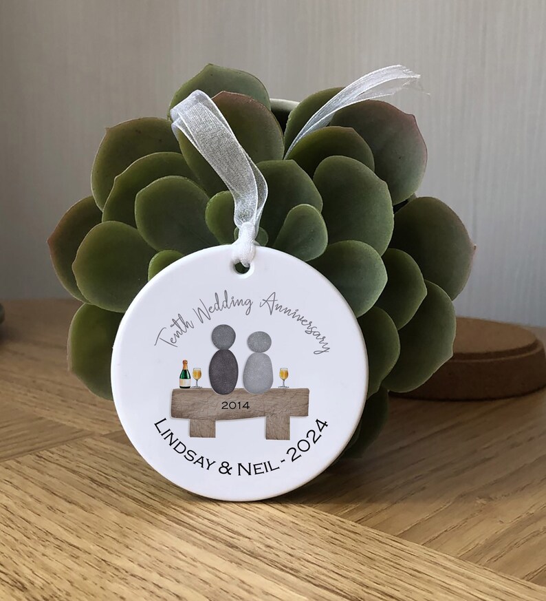 May include: White ceramic ornament with a gray illustration of two people sitting on a bench with a bottle of champagne and glasses. The text "Tenth Wedding Anniversary 2014 - 2024 Lindsay & Neil" is printed on the ornament.