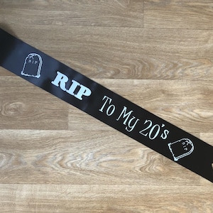 May include: Black satin sash with white text and tombstone graphics. The text reads "RIP To My 20's". A fun accessory for a milestone birthday celebration.