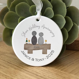 May include: White ceramic ornament with a silver wedding anniversary design. It features two gray pebble figures sitting on a wooden bench with a bottle of champagne and glasses. The text reads "Silver Wedding Anniversary" and "Janice & Tony - 2025".