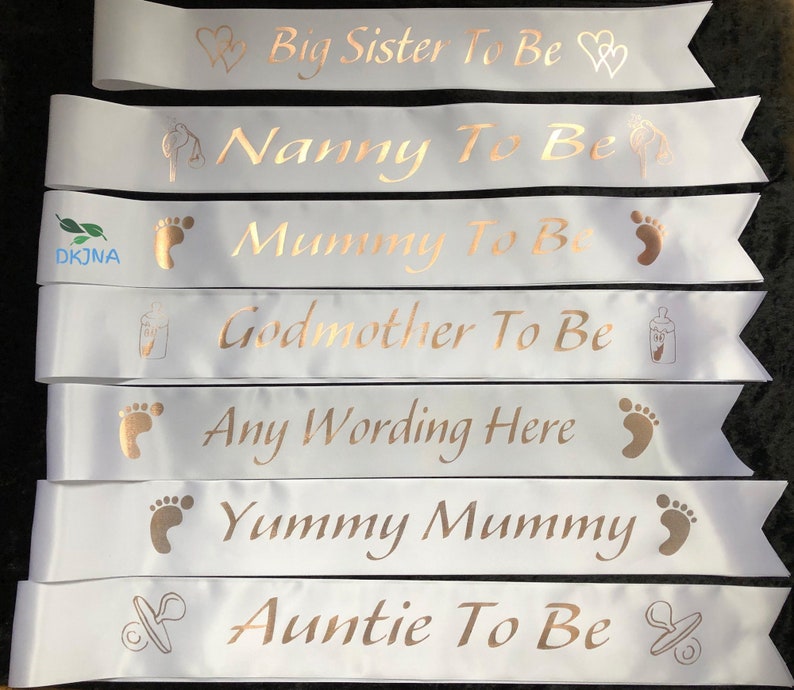 May include: Seven white satin sashes with gold foil lettering and icons. The sashes read "Big Sister To Be", "Nanny To Be", "Mummy To Be", "Godmother To Be", "Any Wording Here", "Yummy Mummy", and "Auntie To Be".