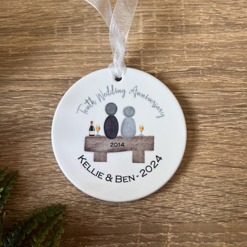 May include: White ceramic ornament with a design of two figures sitting on a bench with a bottle of wine and glasses. The text "Tenth Wedding Anniversary 2014 Kellie & Ben - 2024" is printed on the ornament.
