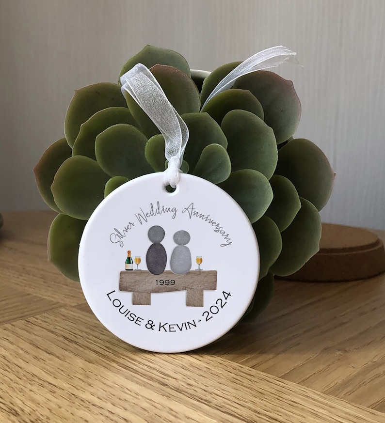 May include: White ceramic ornament with a silver wedding anniversary design. It features two gray figures sitting on a wooden bench with champagne glasses and the text "Silver Wedding Anniversary 1999 Louise & Kevin - 2024".
