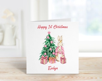 May include: A white card with a pink and green watercolor illustration of a rabbit standing next to a Christmas tree with pink ornaments and presents. The text "Happy 1st Christmas Evelyn" is printed on the card.