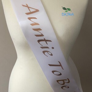 May include: White satin sash with gold lettering that reads "Auntie To Be" and a gold pacifier graphic.