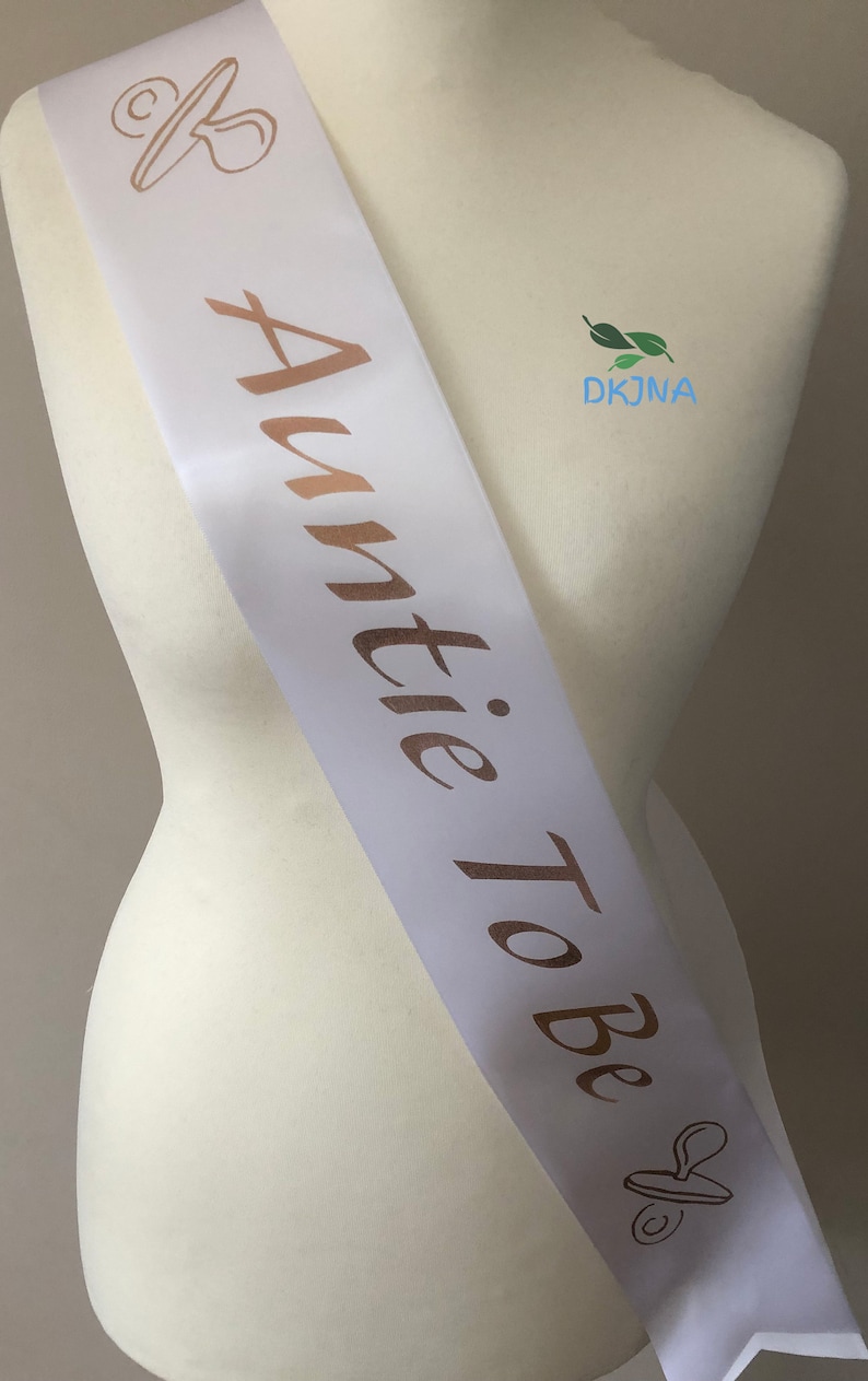 May include: White satin sash with gold lettering that reads "Auntie To Be" and a gold pacifier graphic.