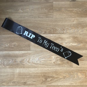 May include: Black satin sash with white text and tombstone graphics. The text reads "RIP To My Teen's". The sash is laid out on a wooden surface.
