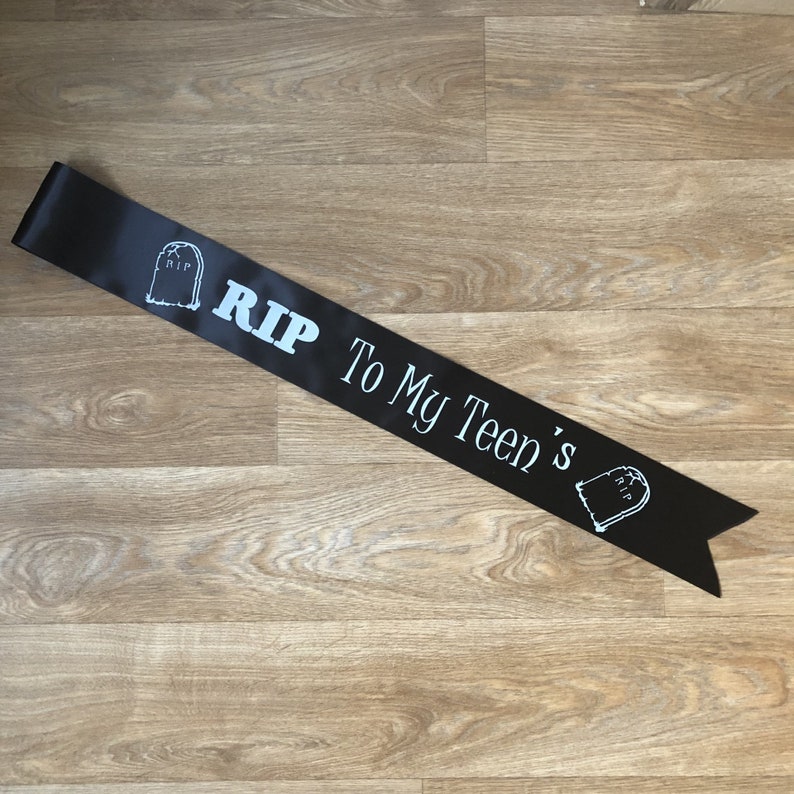 May include: Black satin sash with white text and tombstone graphics. The text reads "RIP To My Teen's". The sash is laid out on a wooden surface.