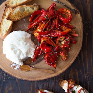 Może przedstawiać: Drewniany talerz do serwowania z wyborem jedzenia. Zawiera tosty, dużą porcję białego sera i stos pieczonej czerwonej papryki. Ozdobione ziołami.