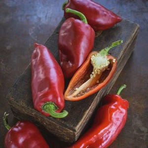 Może przedstawiać: Kilka dojrzałych, czerwonych papryk, niektóre całe, inne przekrojone, ukazujące nasiona. Ułożone na zwietrzałej drewnianej desce, na ciemnym, teksturowanym tle. Obraz skupia się na świeżych produktach.