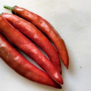 Może przedstawiać: Cztery czerwone papryczki chili z zielonymi łodygami ułożone na białej powierzchni. Papryczki mają gładką, błyszczącą teksturę i różne odcienie czerwieni. Zdjęcie jest zbliżeniem.