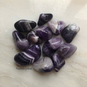 Peut inclure: Une collection de 10 pierres d'améthyste roulées avec des rayures blanches et violettes. Les pierres sont lisses et polies, et elles ont une variété de formes et de tailles.