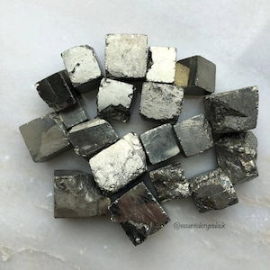 May include: A collection of 15 small, cubic, pyrite crystals. The crystals are a metallic silver colour with a slightly rough texture. The crystals are arranged in a loose pile on a white surface.