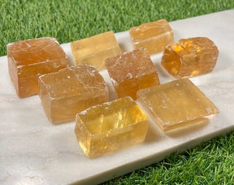 May include: A collection of nine golden selenite crystals, each with a unique shape and size, displayed on a white surface. The crystals are translucent and have a slight yellow hue.