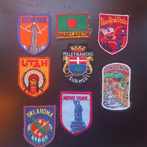 Può includere: Una collezione di toppe ricamate raffiguranti vari luoghi. Le toppe includono "New York" con un grattacielo, "Bangladesh" con una bandiera, "New York State" con un paesaggio urbano, "Utah" con un nativo americano, "Villefranche Sur-Mer", "Kongsvinger", "Oklahoma" e "New York" con la Statua della Libertà.