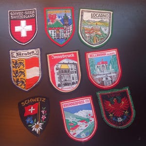 Puede incluir: Una colección de parches bordados con diversos diseños y texto. Los parches muestran imágenes de edificios, paisajes y banderas, con texto como "Suiza", "Innsbruck" y "Tirol". Los parches tienen forma de escudo con bordes coloridos.