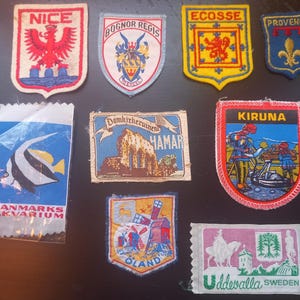 May include: A collection of vintage travel patches and a Danmarks Akvarium label. The patches feature embroidered designs with text like "NICE", "ECOSSE", "PROVENCE", "KIRUNA", "OLAND", and "Uddevalla SWEDEN". The patches are in various shapes and colors.