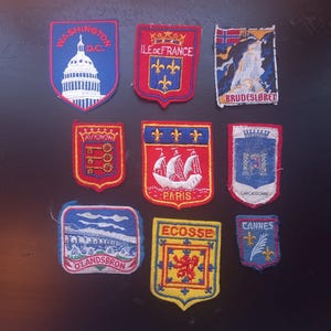 Può includere: Una collezione di toppe ricamate raffiguranti vari luoghi. Le toppe includono emblemi di Parigi, Cannes e altre città europee. Le toppe sono in vari colori e forme, con dettagli ricamati.