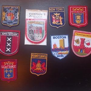 May include: A collection of vintage souvenir badges featuring embroidered designs. The badges showcase various locations, including Great Britain, New York, Boston, and Amsterdam. Each badge has a unique shape and color scheme, with text indicating the place.