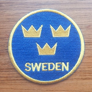 May include: A circular embroidered patch featuring a vibrant blue background with a gold border. The design includes three gold crowns arranged in a triangular pattern above the word SWEDEN, which is stitched in gold capital letters.