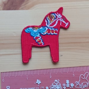 May include: Red felt Dala horse applique with blue, white and yellow embroidered details. The horse is standing on its four legs and has a long, flowing mane.