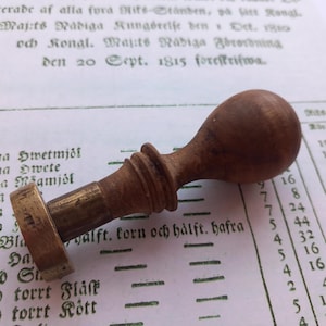 May include: A wooden and brass seal with a round handle and a circular base. The seal is used to stamp documents and is a vintage item.