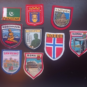 Op de afbeelding: Een verzameling geborduurde patches met verschillende stads- en landemblemen. Patches zijn onder meer "Pakistan", "Stockholm", "St. Paul's Cathedral", "København", "Delfi", "Norge", "Taormina", "San Remo Casino" en "Roma". Elke patch heeft een uniek ontwerp en kleurenschema.