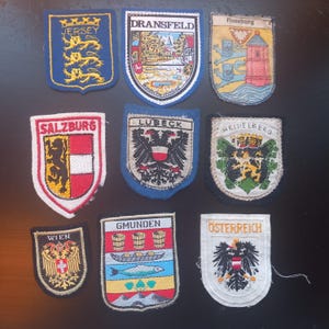May include: A collection of nine embroidered patches featuring various city and country crests. The patches are in various shapes and sizes, with text such as "Jersey", "Dransfeld", and "Salzburg". The patches are in various colors, including blue, red, and yellow.