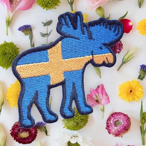 Op de afbeelding: Geborduurde patch in de vorm van een eland, met de Zweedse vlag. De eland is blauw met een gouden kruis. De patch is omgeven door kleurrijke bloemen, waaronder roze, gele en witte bloemen.