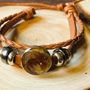 May include: A brown leather bracelet with a braided design and a round, resin-filled charm. The charm has a brown and gold swirl pattern. The bracelet has two silver metal beads.