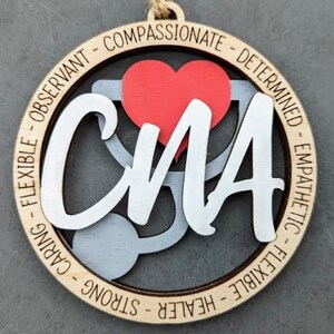 May include: A wooden ornament with a stethoscope and heart design. The words "CMA" are written in white on the ornament. The words "Flexible, Observant, Compassionate, Determined, Empathetic, Flexible, Healer, Strong, Caring" are written around the edge of the ornament.