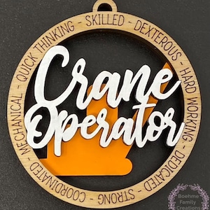 May include: A wooden ornament featuring a white silhouette of a crane operator and the words "Crane Operator" in white lettering. The ornament is decorated with a brown wooden ring with the words "Quick Thinking - Skilled - Dexterous - Hard Working - Dedicated - Strong - Coordinated" in maroon lettering.