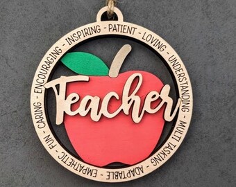 Può includere: Ciondolo in legno a forma di mela rossa con una foglia verde. La parola "Teacher" è scritta in corsivo sulla mela. Il ciondolo è circondato da un cerchio con le parole "Encouraging, Inspiring, Patient, Loving, Understanding, Multi-Tasking, Adaptable, Empathetic, Fun, Caring" scritte intorno al bordo.