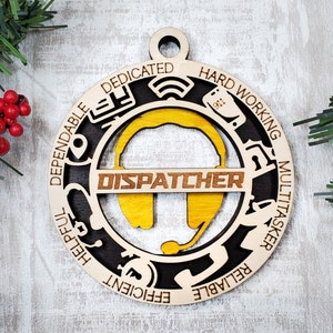 May include: A wooden Christmas ornament with a yellow headset and the word "DISPATCHER" in the center. The ornament is surrounded by a black circle with the words "Dependable, Dedicated, Hard Working, Multitasker, Reliable, Efficient, Helpful" written around the edge.