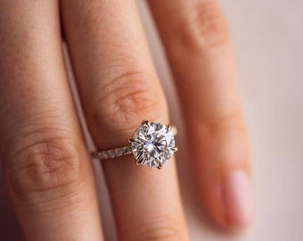 May include: A close-up of a diamond ring with a large, round-cut center stone. The ring has a gold setting and a band adorned with smaller diamonds. The ring is on a finger.