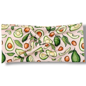 May include: A light pink headband with a repeating pattern of avocados, avocado slices, leaves, and avocado pits. The avocados are green and the pits are brown. The headband is twisted at the top.