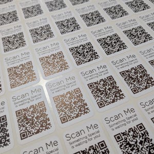 May include: A collection of white stickers, each featuring a QR code. The stickers display the text "Scan Me" and "Something Special is waiting for you!". The QR codes are printed in black and brown.