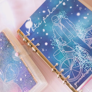 Può includere: Un raccoglitore blu e verde a tema galassia con un contorno bianco di una persona che va in bicicletta. Il raccoglitore ha un meccanismo ad anelli dorati e il testo "let's palace" e "goroceewintershop" stampato sulla copertina.