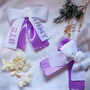 Può includere: Due fiocchi per capelli bianchi e viola con le parole "BTS" e "ARMY" stampate sui nastri. I fiocchi sono su una superficie di raso bianco, con fiori secchi e verde.