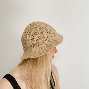 A blond woman is wearing a beige crochet bucket hat. A hat is made from granny squares with a single crochet brim. 
A woman is wearing a black tank top and there is a white background.