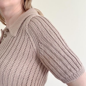 May include: A light brown, short-sleeved knit polo shirt with a ribbed texture. The shirt features a collared neckline and button details. The sleeves are a shorter length, and the overall style is classic and versatile.