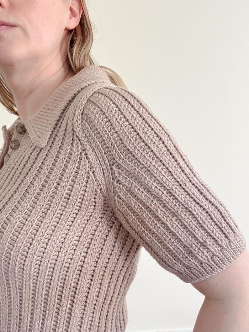 May include: A light brown, short-sleeved knit polo shirt with a ribbed texture. The shirt features a collared neckline and button details. The sleeves are a shorter length, and the overall style is classic and versatile.