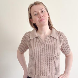 May include: A tan-colored, short-sleeved knit polo shirt with a collared neckline and three buttons. The shirt has a ribbed texture and a V-neck design. The model is wearing black pants.